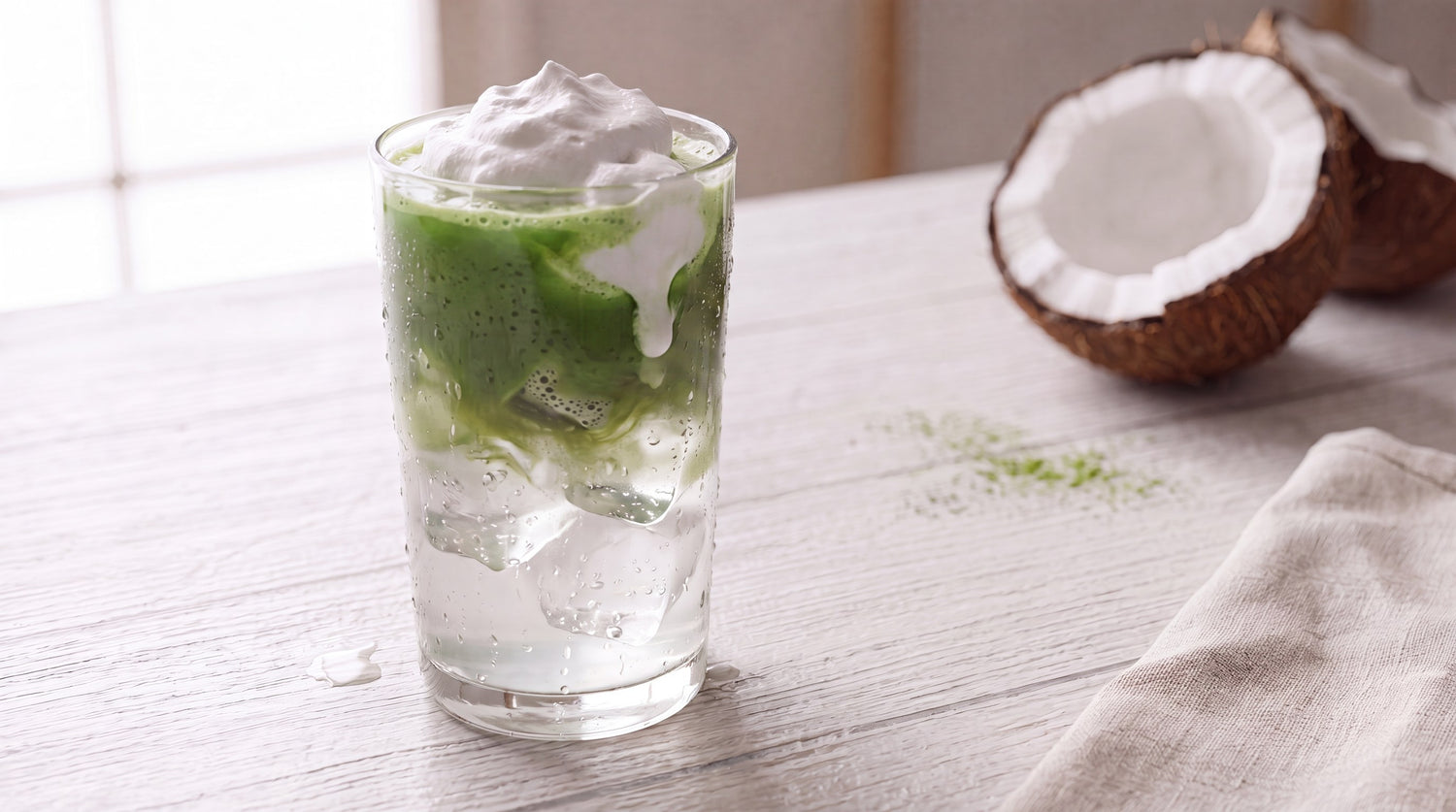 Coconut matcha cloud in a tall clear glass with vivid natural green matcha from Japan layered over ice and chilled coconut water, topped with a whipped coconut cream cloud, beside a fresh cracked coconut on a light wood table